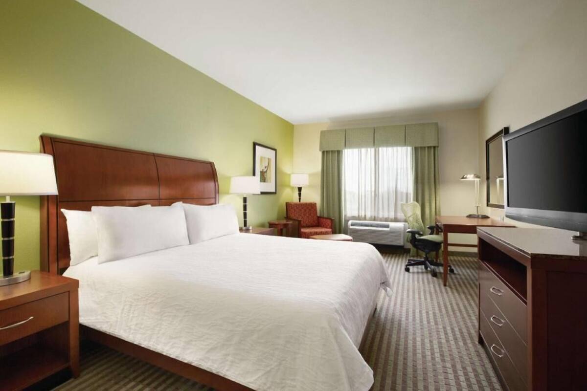 A hotel room with a bed, two bedside tables, a desk with a chair, a television, and a window with curtains.