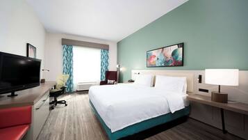 Una habitación de hotel con una cama, un escritorio con televisor, una silla, una lámpara de escritorio y un cuadro en la pared.
