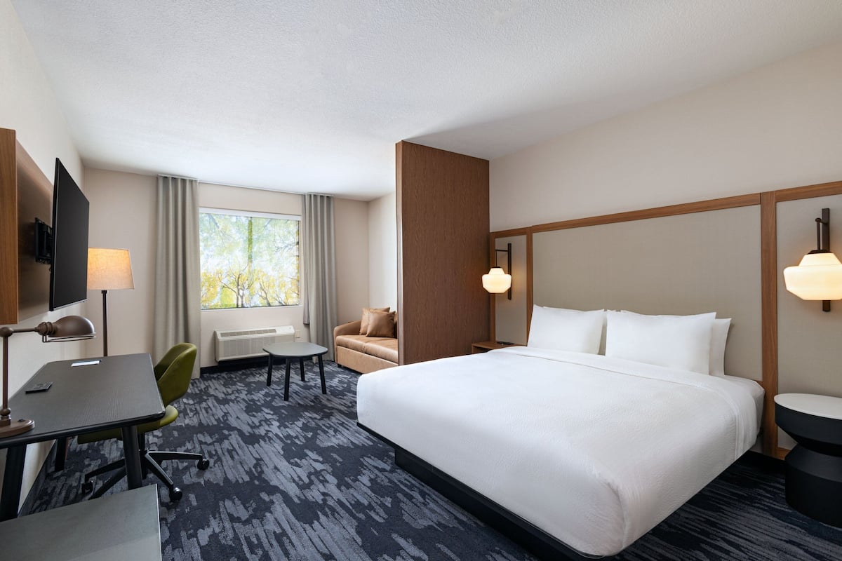 A hotel room with a large bed, a desk, a chair, a small table, a TV, and a window with curtains.