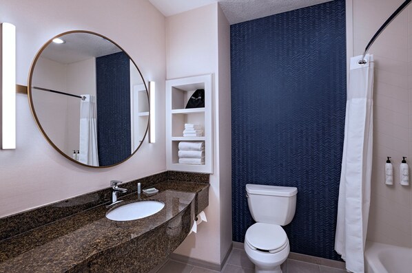 Une salle de bain moderne avec un comptoir en granit, un miroir rond, un lavabo blanc, des toilettes et un rideau de douche.