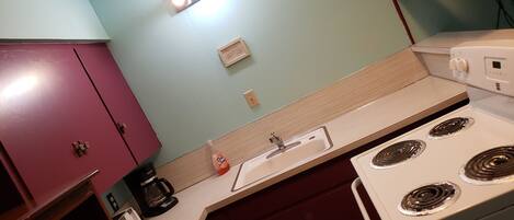 A kitchen with a white stove, a sink, and a coffee maker. There is a cabinet with a red door and a light switch on the wall.