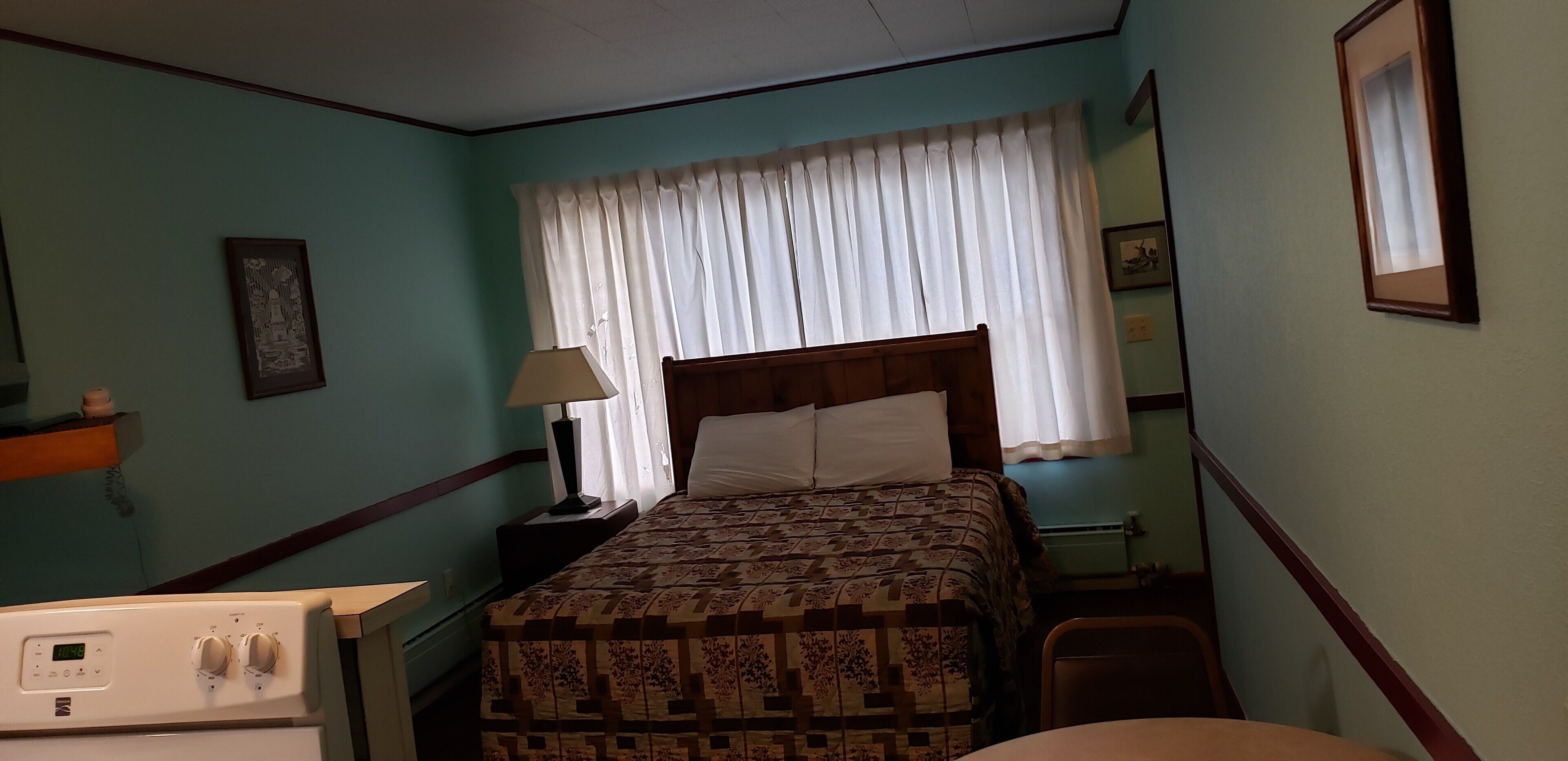 Un dormitorio con cama, mesita de noche con lámpara, una silla, una pequeña mesa y un calentador fijado a la pared.