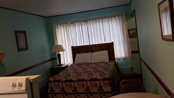Un dormitorio con cama, mesita de noche con lámpara, una silla, una pequeña mesa y un calentador fijado a la pared.