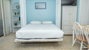 Ein Schlafzimmer mit einem Bett, einem Fernseher auf einem Schrank, einem Sessel und einem Regal mit Dekoration.