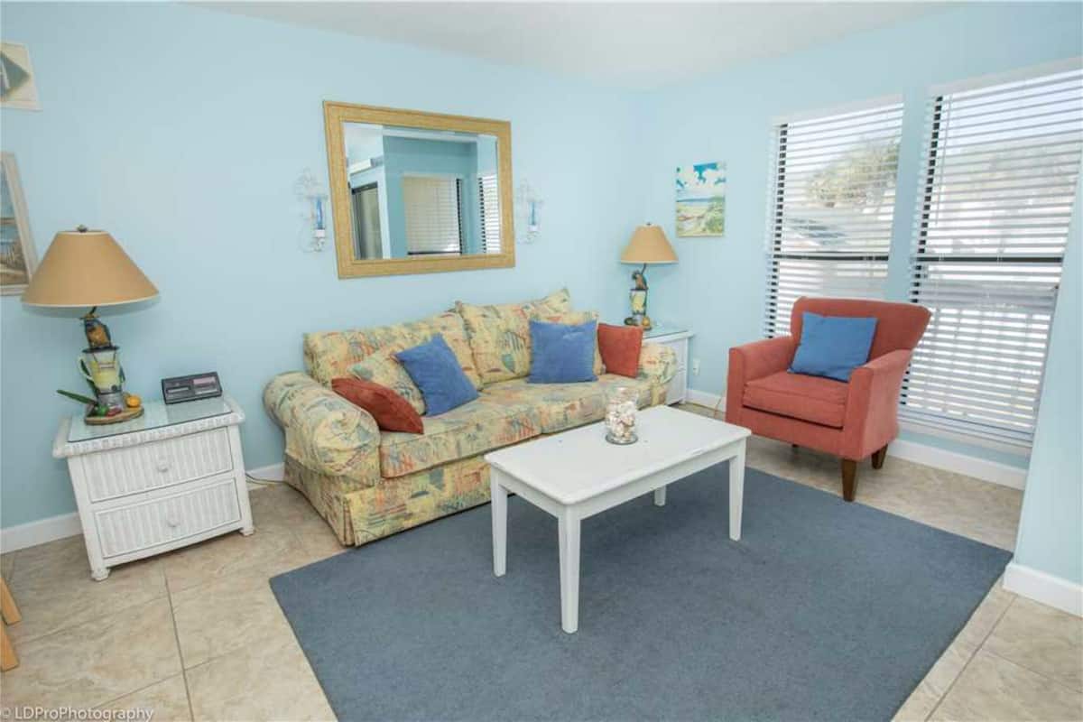 A living room with a patterned sofa, a white coffee table, a red armchair, and a small white side table with a lamp.