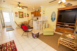 A living room with a TV, a dining area with chairs, a kitchen with cabinets, and a ceiling fan.