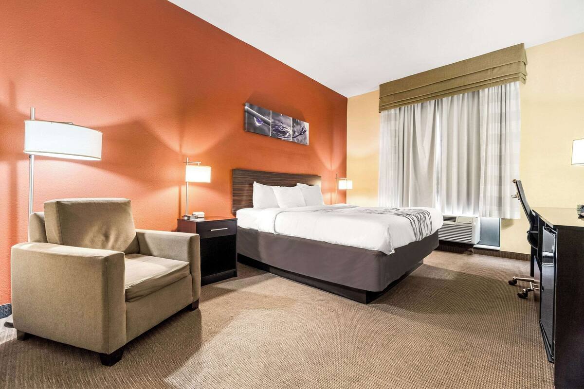Habitación de hotel con cama, una silla, escritorio y un ventanal con cortinas.