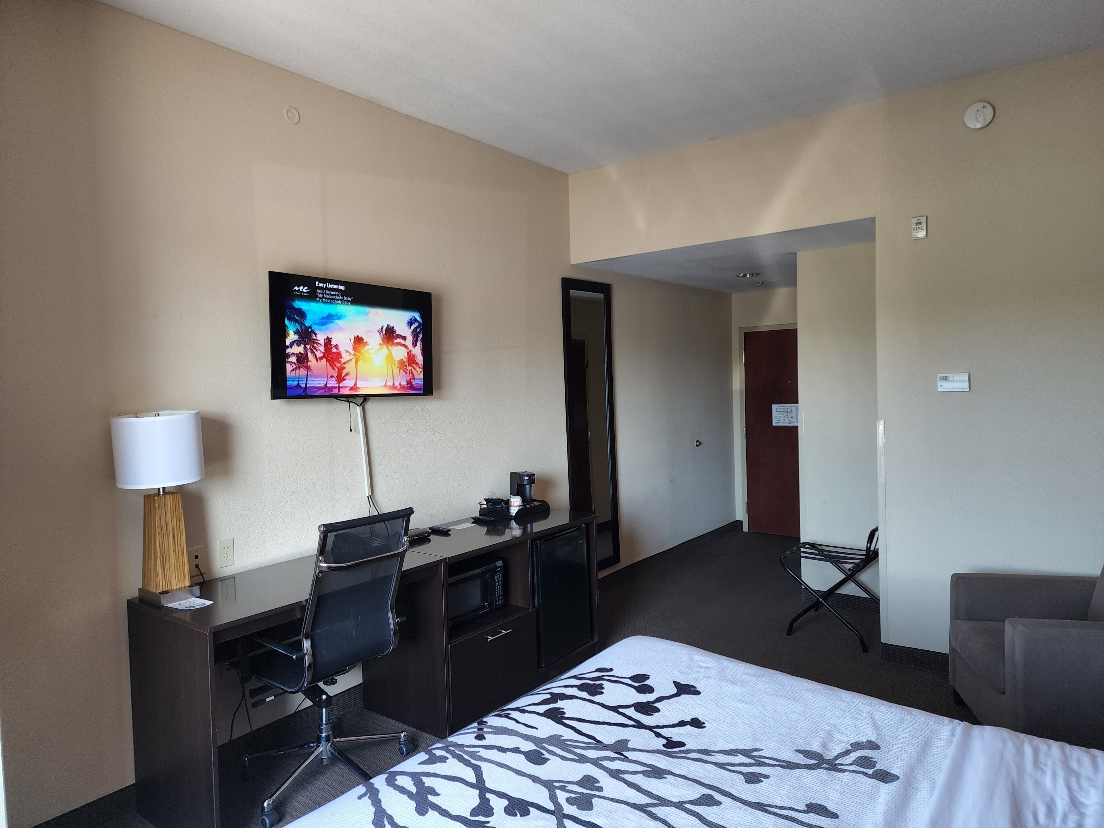 Habitación de hotel con cama, escritorio, silla, televisión, lámpara y vista al pasillo.