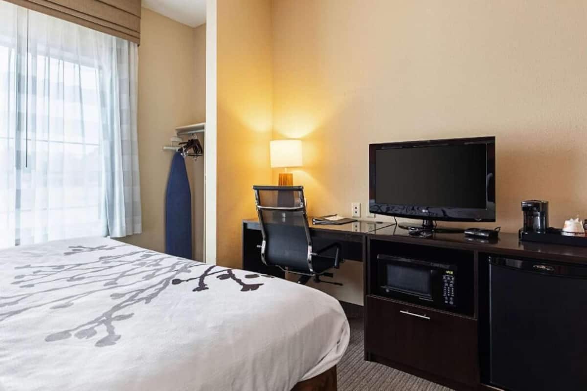 Una habitación de hotel con una cama, un escritorio con televisor, una silla, una lámpara y una cafetera.