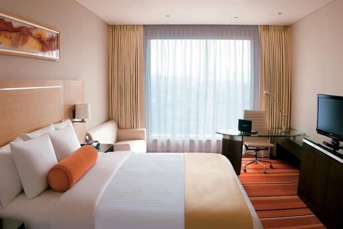 Quarto de hotel com uma cama grande, uma escrivaninha com cadeira, televisão e janela com cortinas.