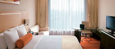 Habitación de hotel con una cama grande, un escritorio con computadora, televisor, una silla y una ventana con cortinas.