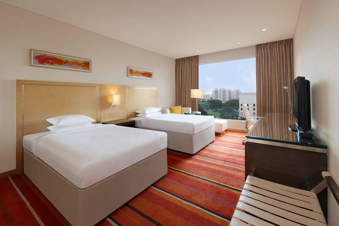 A hotel room with two beds, a desk, a TV, and a large window with a city view.