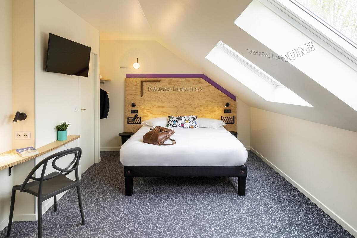 A bedroom with a bed, a desk, a chair, a TV, a window, and a ceiling light.