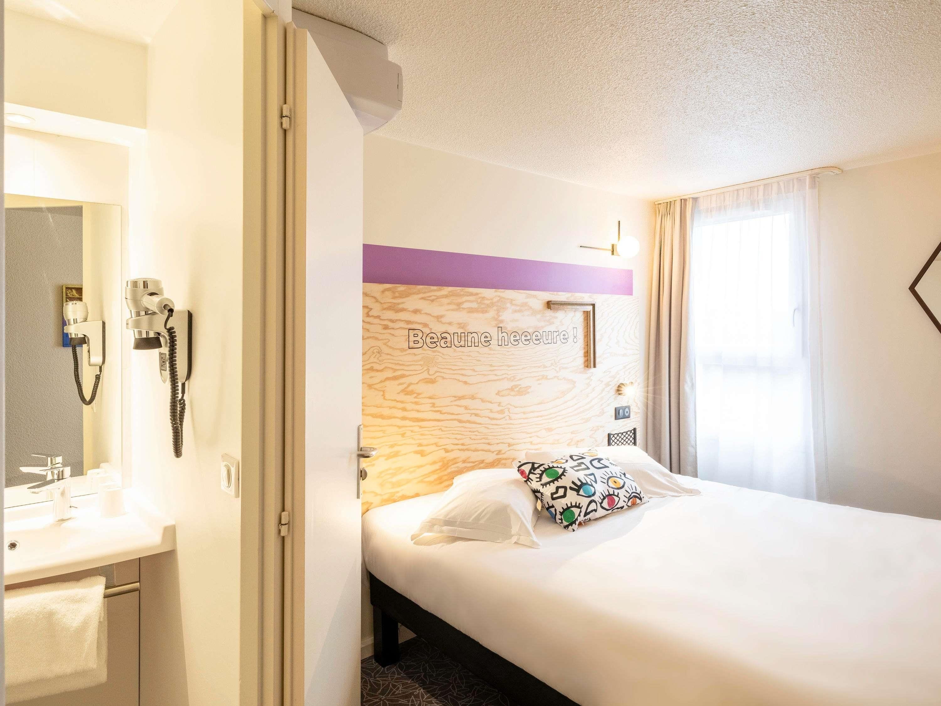 A hotel room with a bed, a bedside table, a mirror, a phone, and a window with curtains.