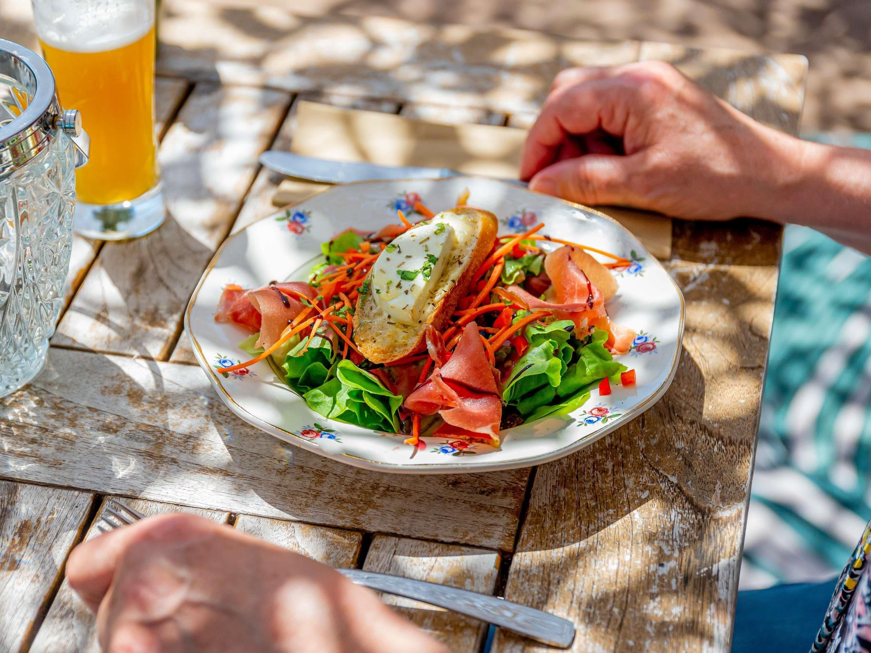 A plate of food with a salad, prosciutto, and a slice of bread, accompanied by a glass of beer on a wooden table.