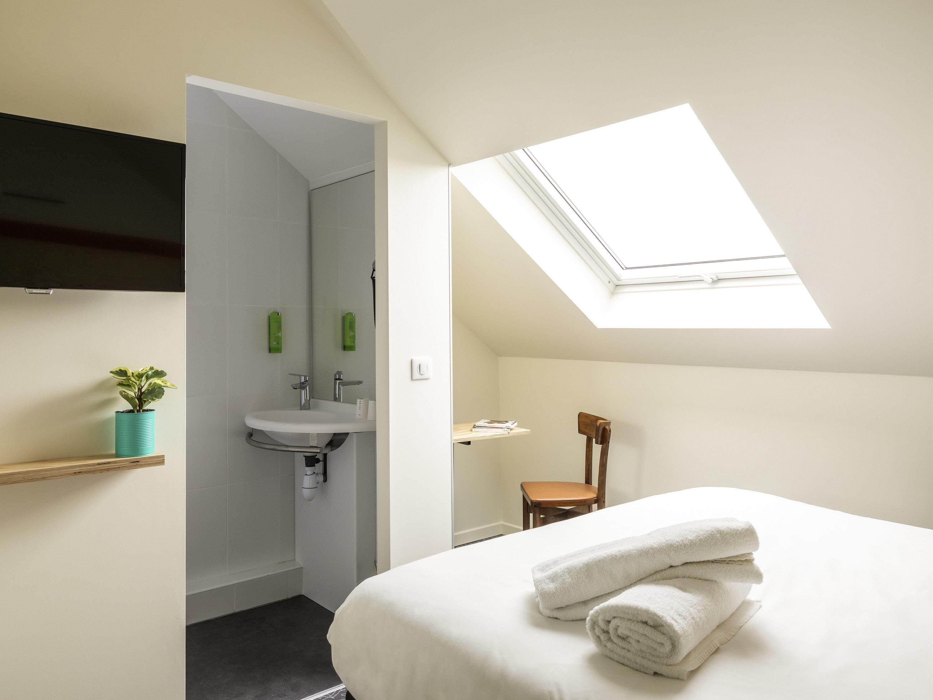 A bedroom with a skylight, a bed with white linens, a bedside table, a chair, and a small bathroom with a sink and mirror.