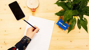 A hand writing on a lined paper with a 'greet' label, a smartphone, a glass, and a potted plant on a wooden surface.
