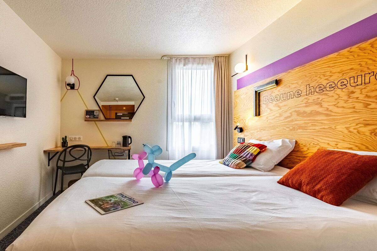 A hotel room with a bed, a desk, a chair, a mirror, and a window with curtains.