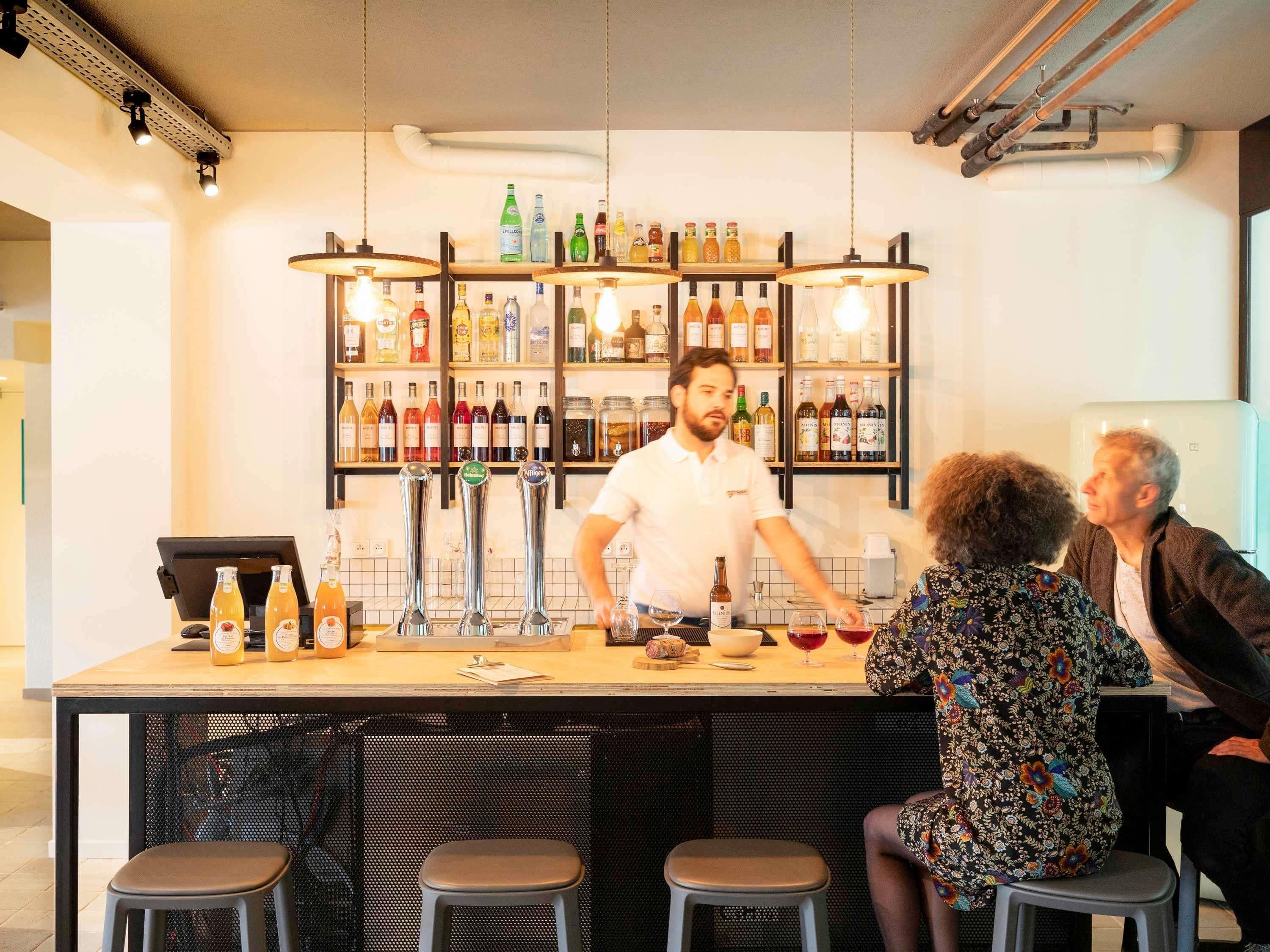 A bar area with a bartender, stools, a counter with bottles, and a shelf with various bottles.