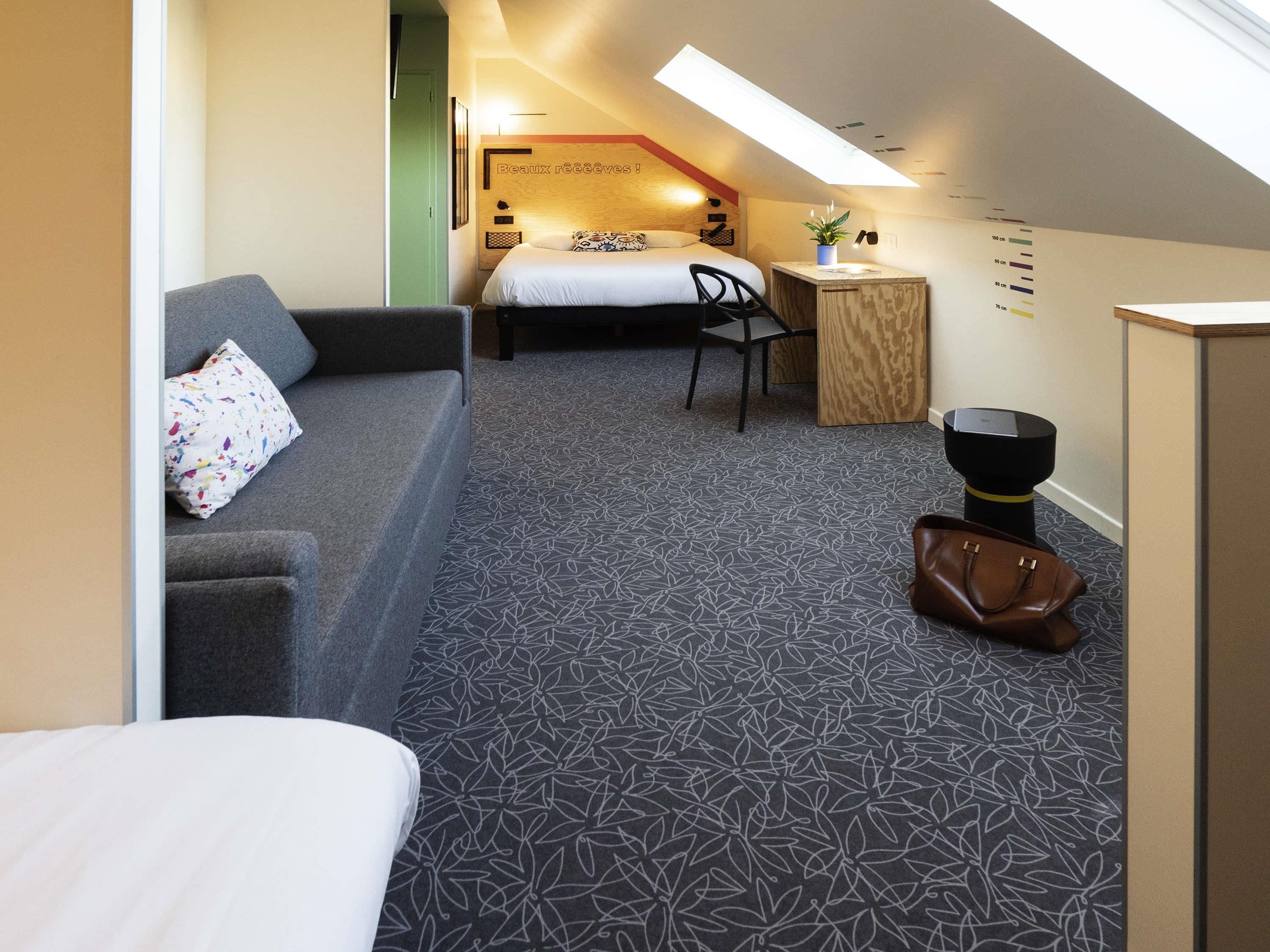 A compact hotel room with a bed, a sofa, a small table, a chair, and a desk.
