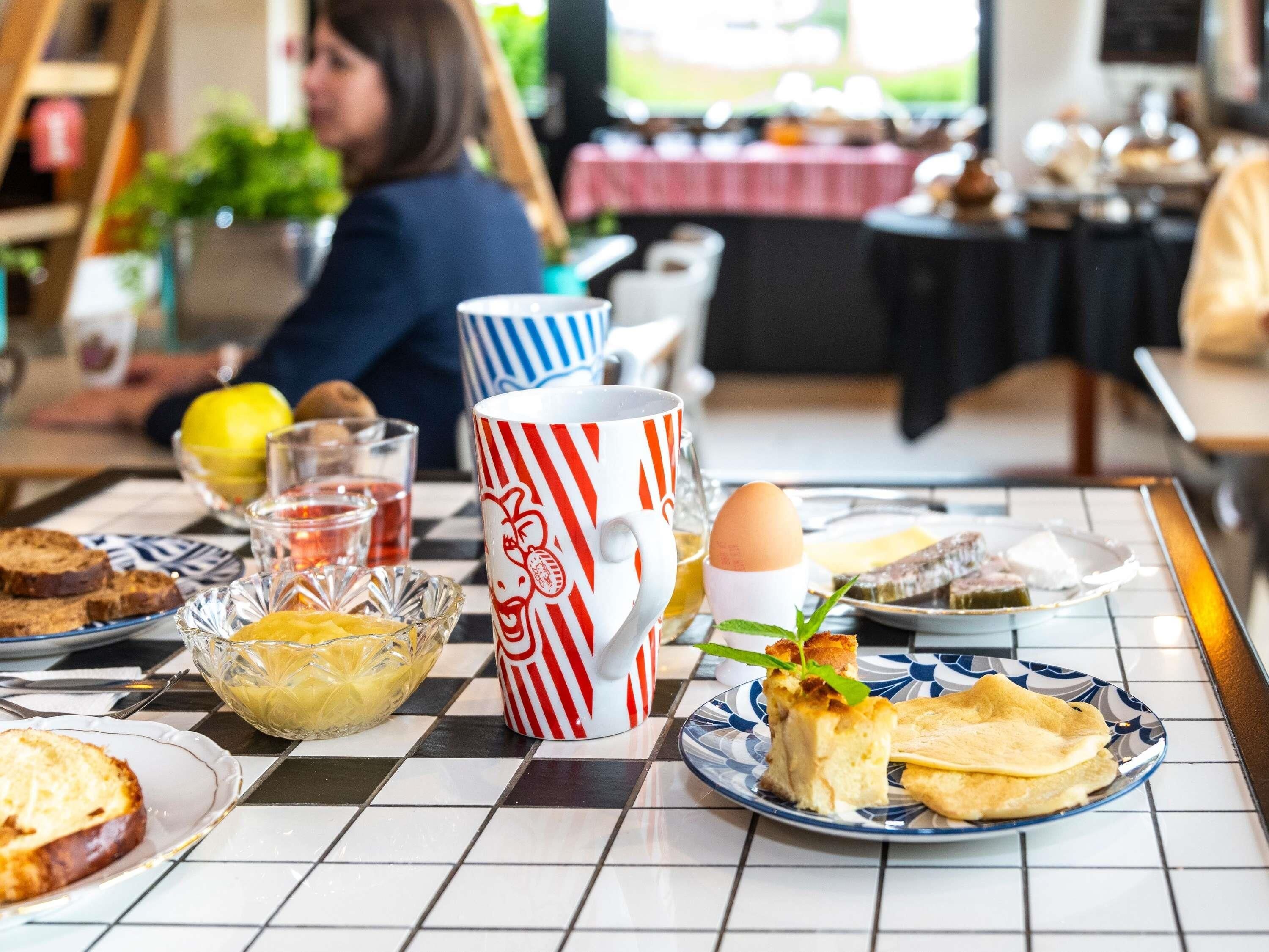Une table mise pour le petit-déjeuner, avec une nappe à carreaux, une tasse rayée, une assiette avec des toasts et une part de gâteau, et un bol de fruits.