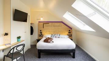 A bedroom with a bed, a desk, a chair, a TV, a window, and a ceiling light.