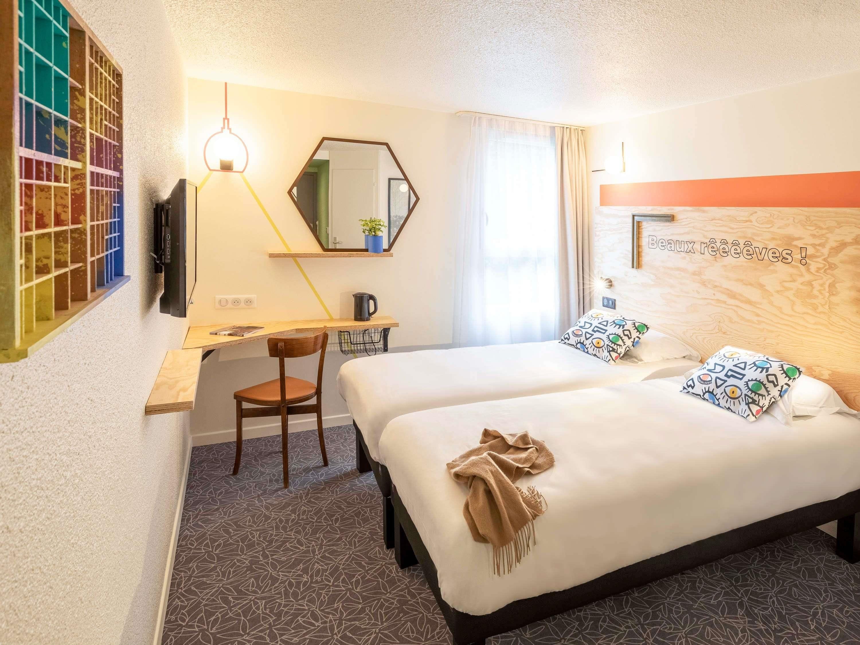 A hotel room with two beds, a desk, a chair, a mirror, a television, and a window with curtains.