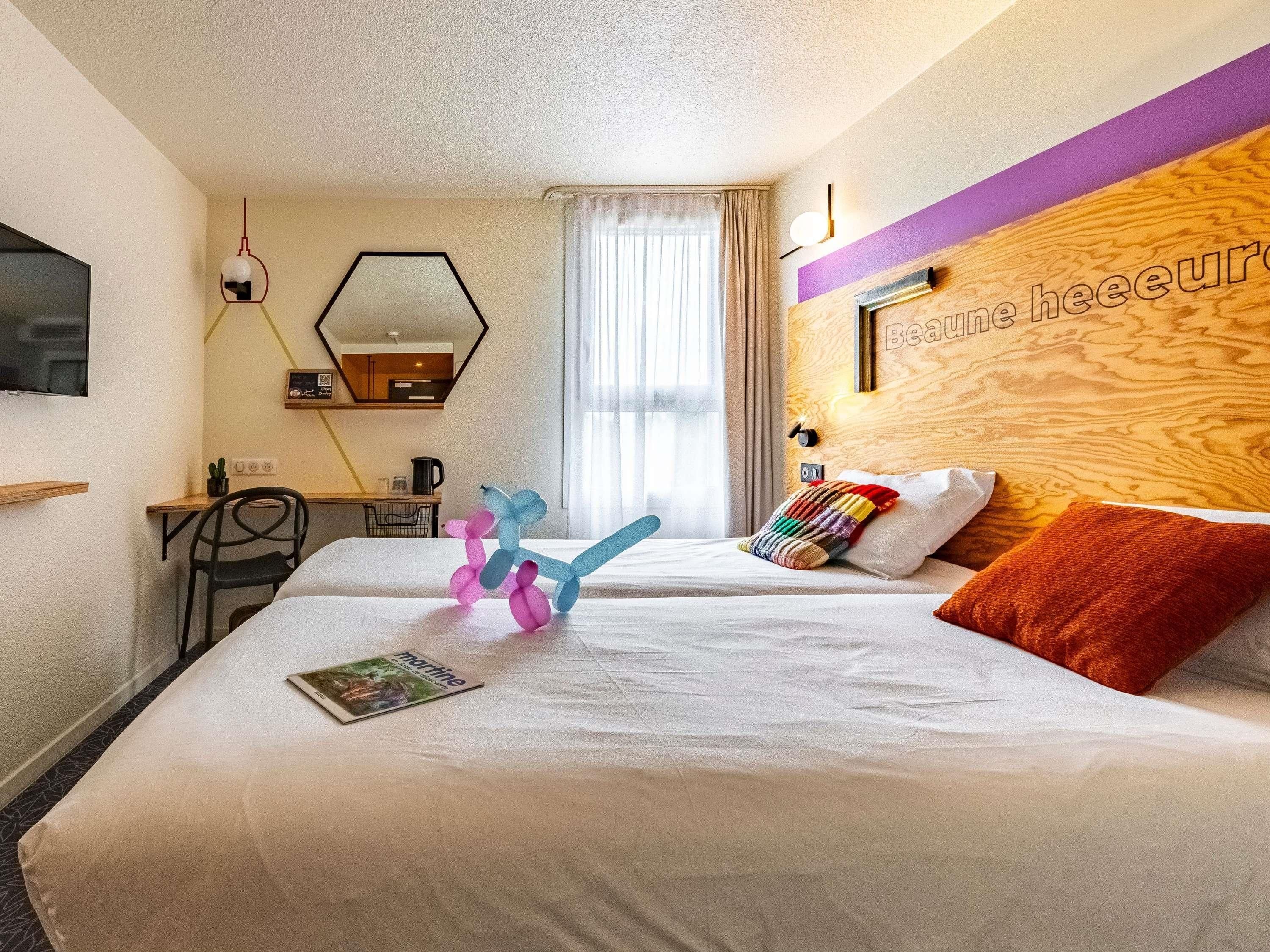 A hotel room with a bed, a desk, a chair, a mirror, and a window with curtains.