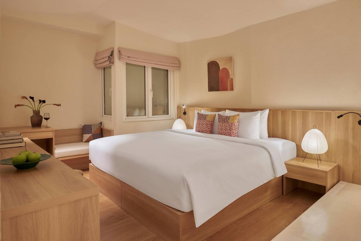 Habitación de hotel impecable con una cama grande, mesitas de noche, un escritorio con un bol de manzanas y una ventana con cortinas.