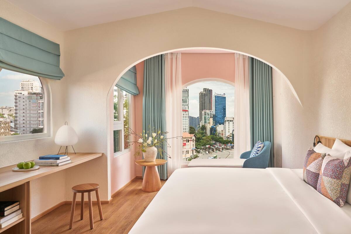 Habitación de hotel con una cama grande, un escritorio con libros, una silla y vistas de la ciudad a través de un ventanal en arco.