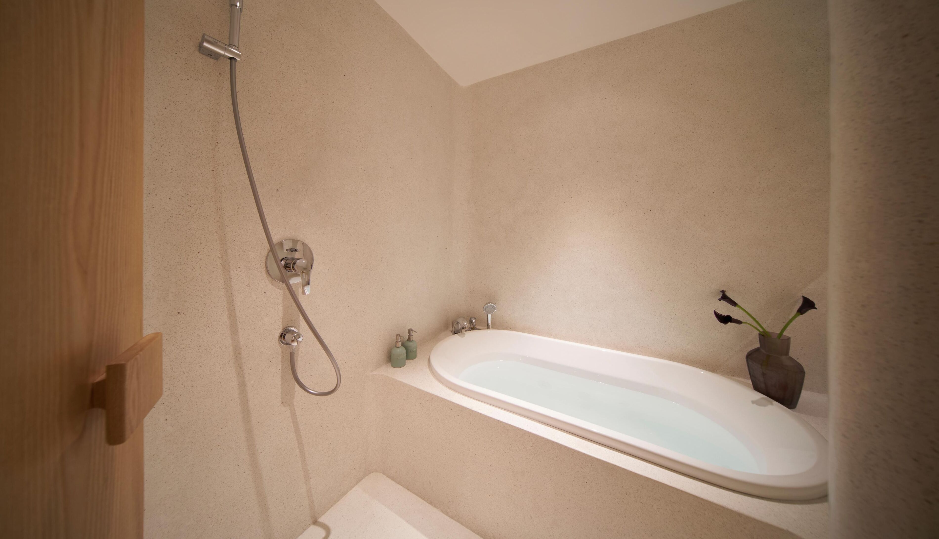 A bathroom with a bathtub, shower, and a small sink. There is a wooden door and a vase with flowers on the sink.
