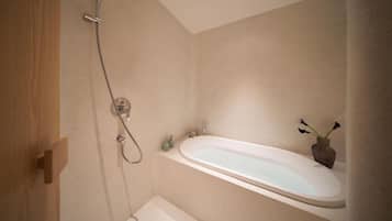A bathroom with a bathtub, shower, and a small sink. There is a wooden door and a vase with flowers on the sink.
