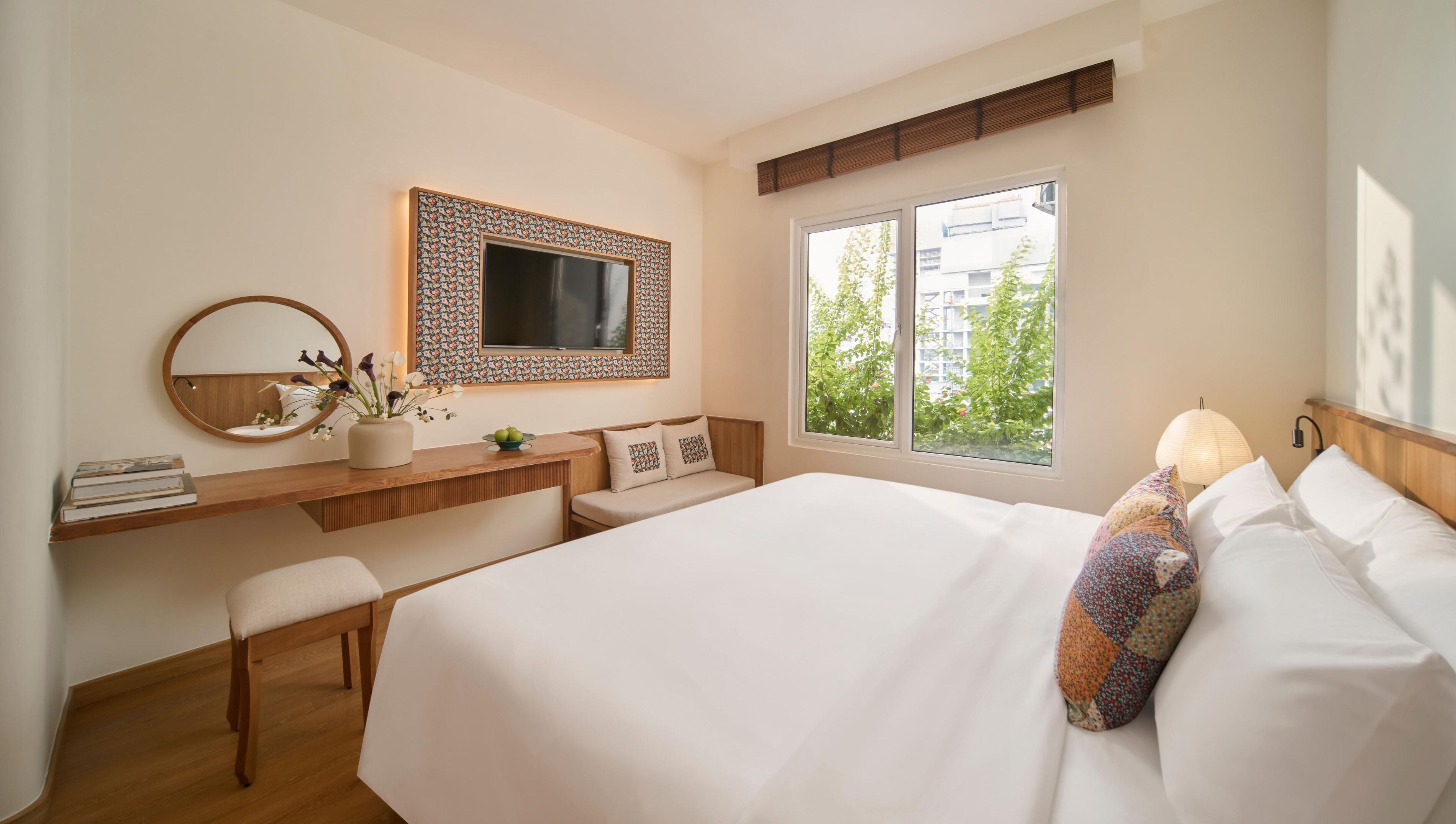 A modern hotel room with a large bed, a bedside table with a mirror, a small desk, and a window with a view of greenery and buildings.