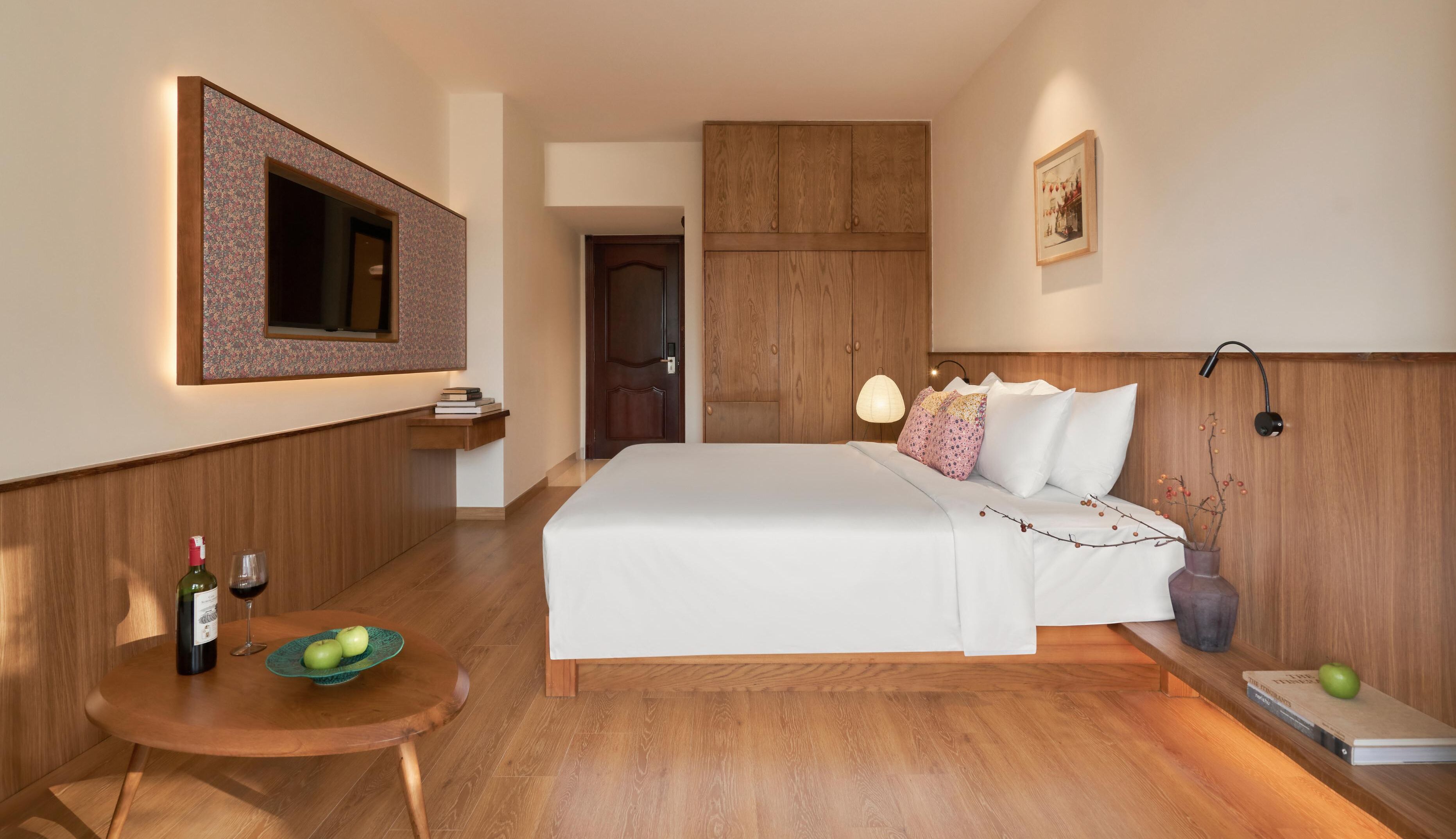 Habitación de hotel con una cama grande, un armario de madera, una mesita con una botella y vasos, y un cuadro en la pared.