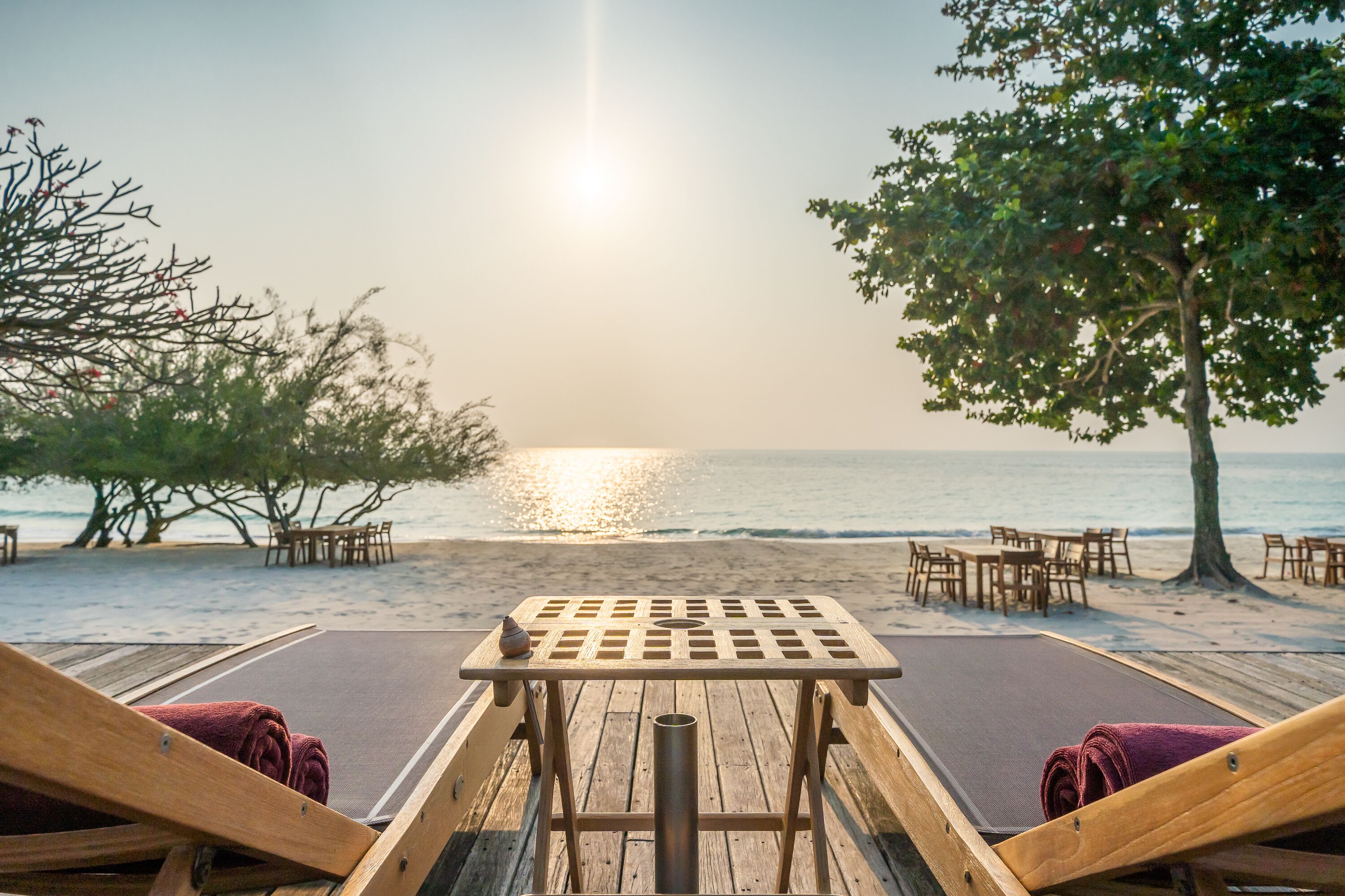 A wooden deck with a table and two lounge chairs, overlooking a beach with a sunset.