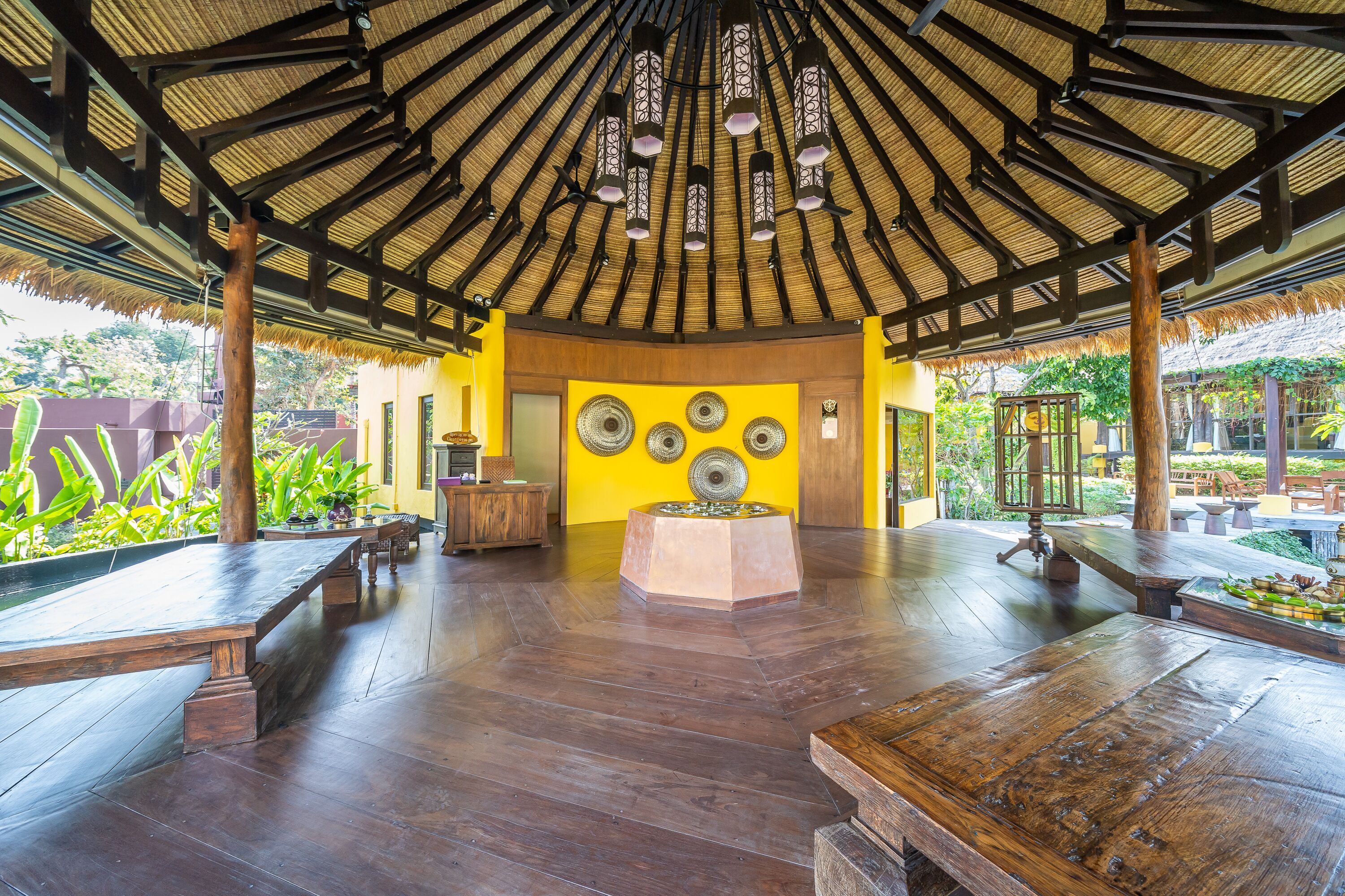A covered outdoor area with wooden benches, a central decorative table, and a thatched roof structure.