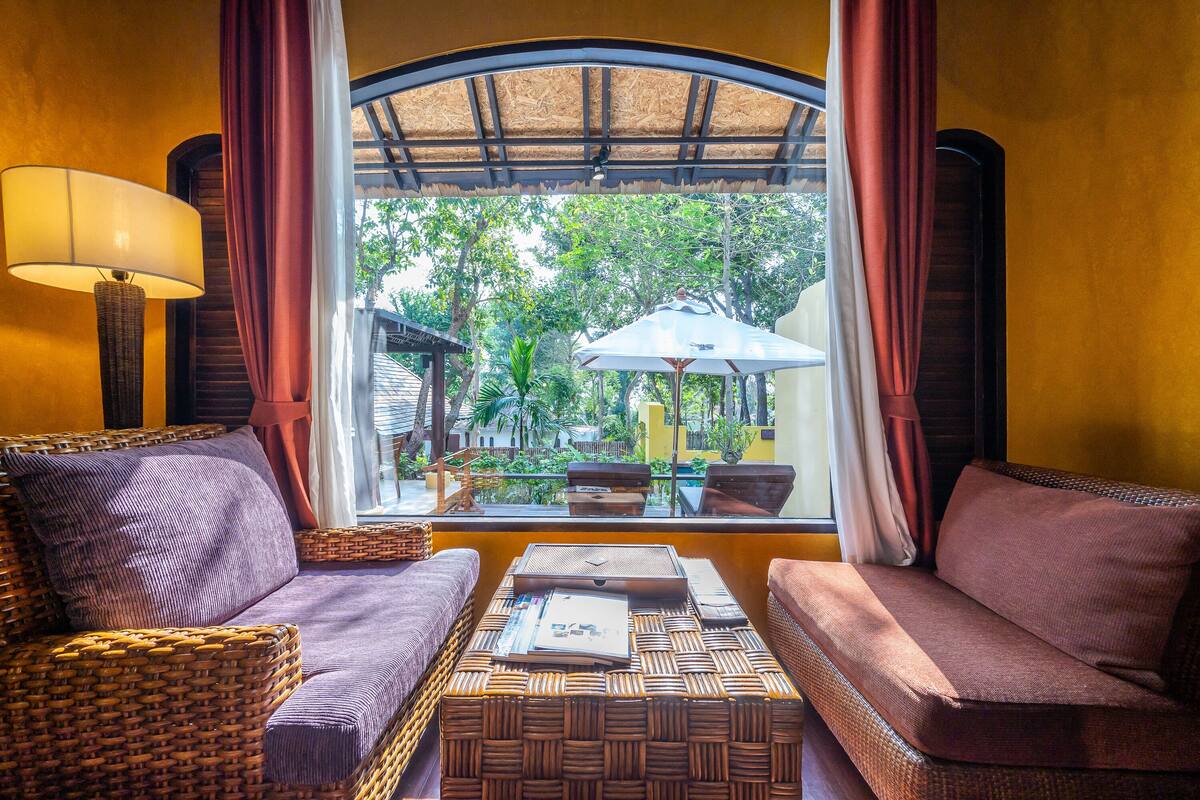 A room with a wicker sofa, a small table with a magazine, and a view of the outdoors through a window with curtains.