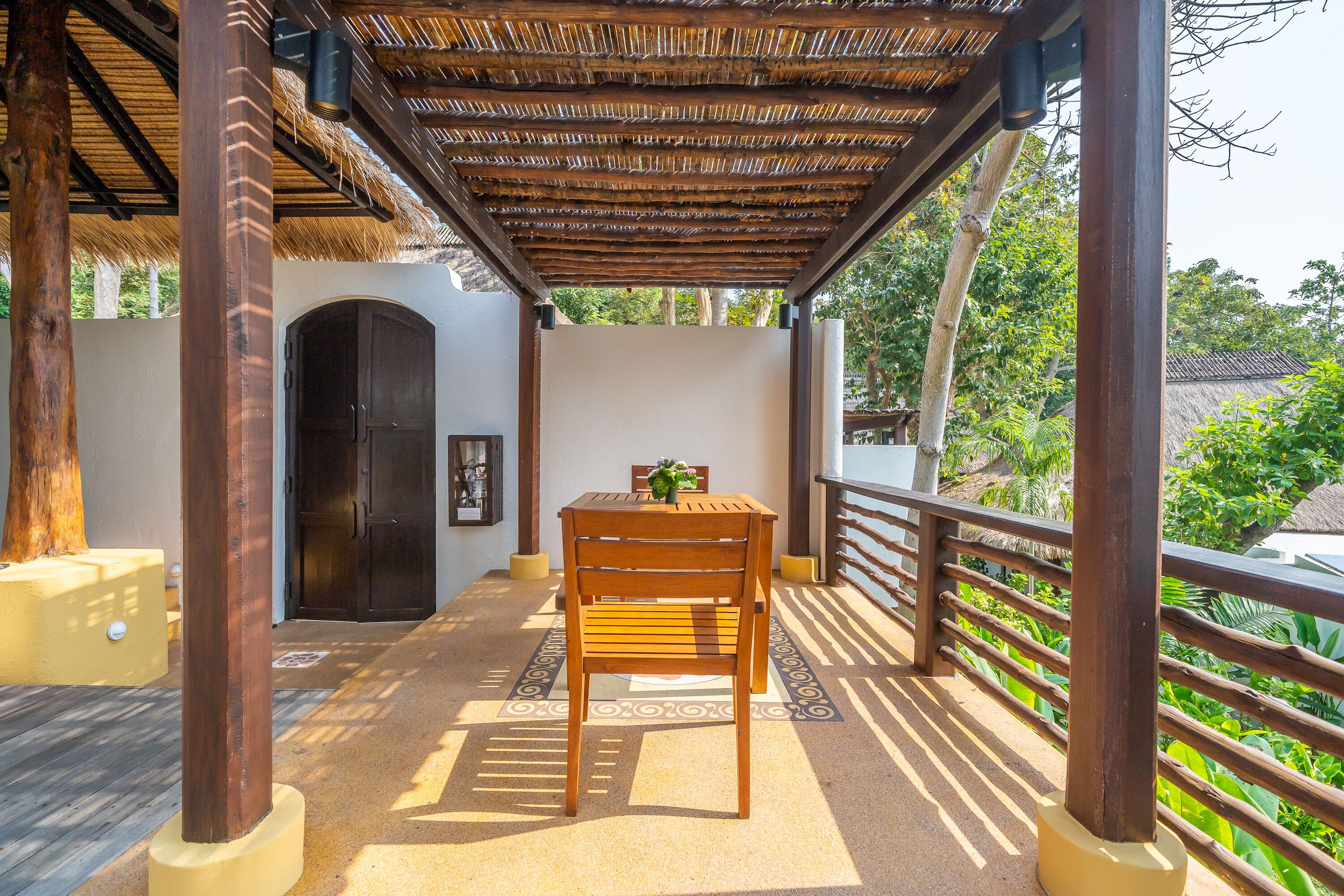 Mesa y silla de madera bajo una estructura de techo de paja, con vistas a follaje y un edificio al fondo.