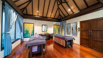 A room with two massage tables, wooden flooring, a ceiling fan, and large windows with curtains.