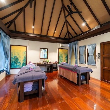 A room with two massage tables, wooden flooring, a ceiling fan, and large windows with curtains.