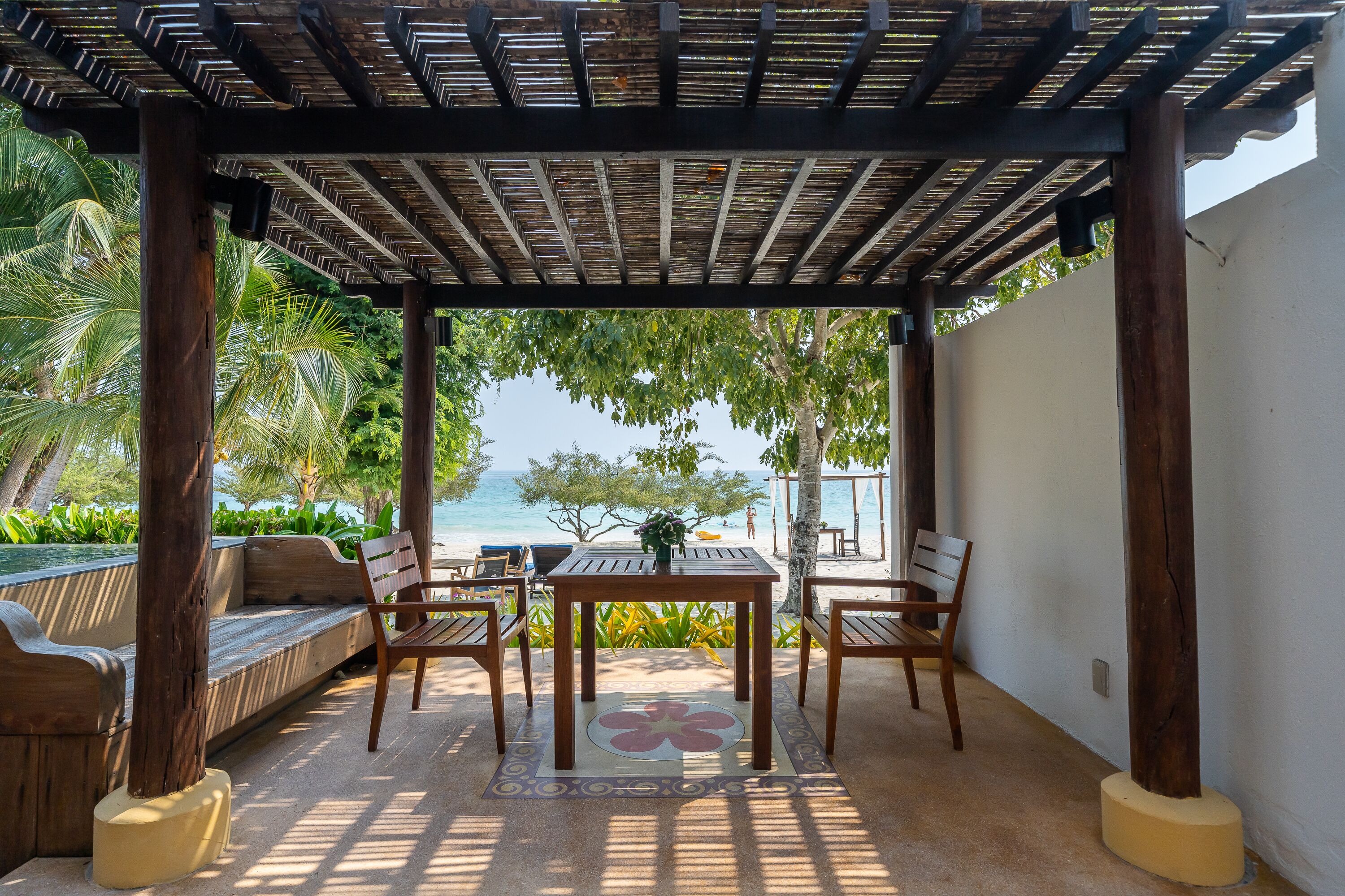 A covered outdoor seating area with wooden furniture and a table, overlooking a beach.
