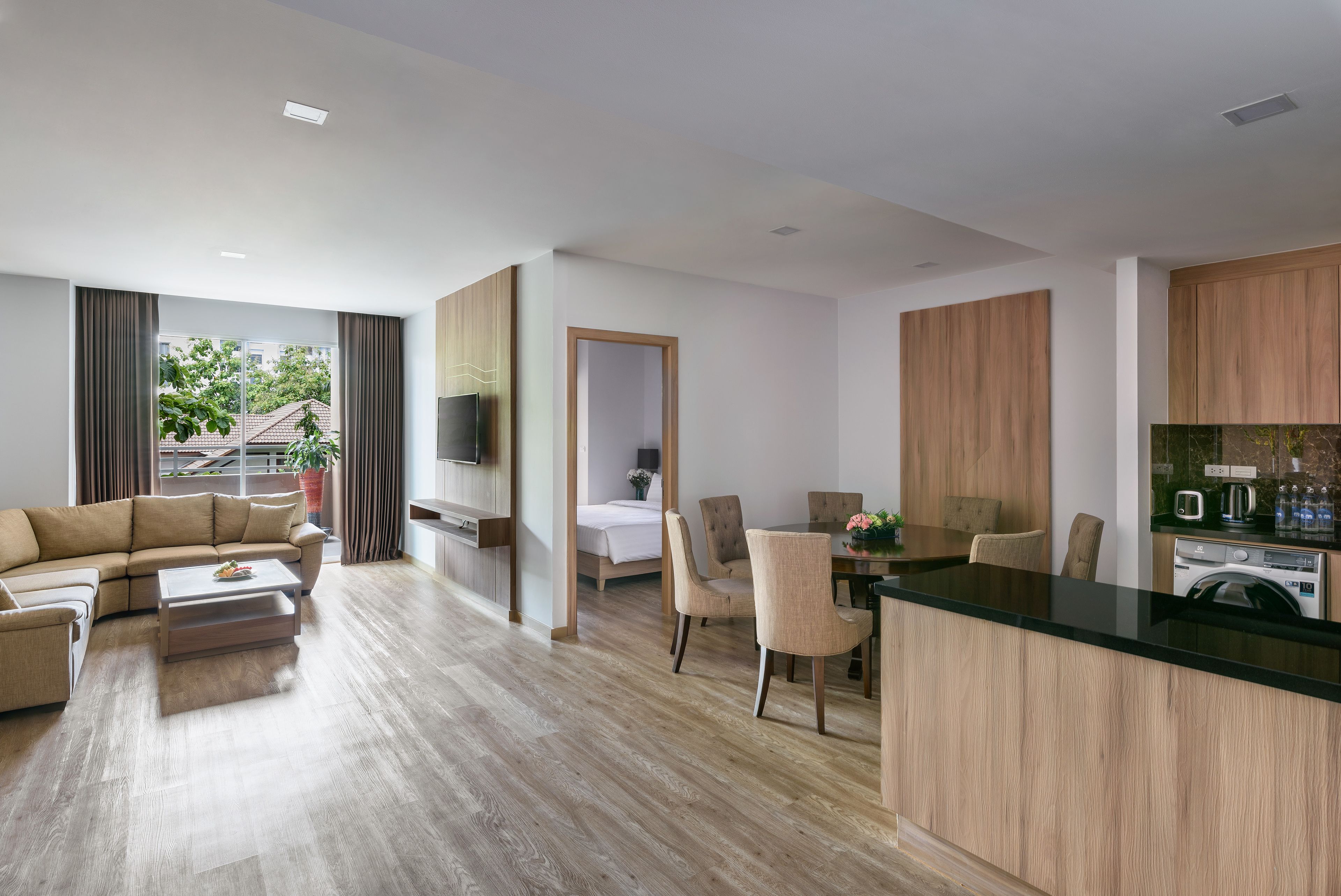 A modern living room with a sofa, coffee table, and dining area. There is a kitchen with wooden cabinets and a countertop. A TV is mounted on the wall, and a window provides a view of the outside.