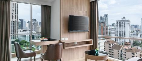 A modern hotel room with a large window, a flat-screen TV mounted on the wall, a dining table with chairs, and a small round table with a plant.