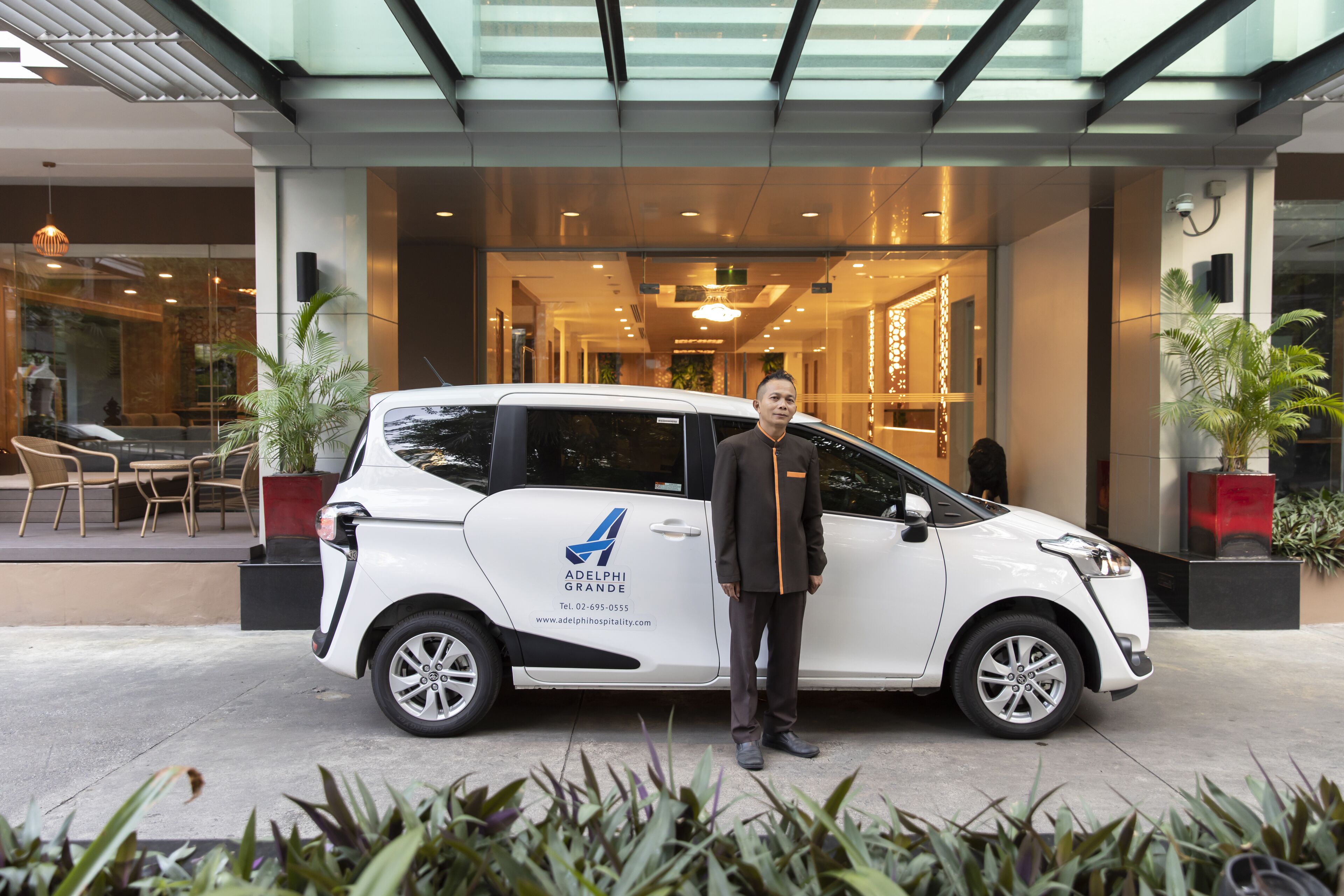 A white car parked in front of a building with a glass entrance, a man standing beside it, and indoor seating visible through the glass.