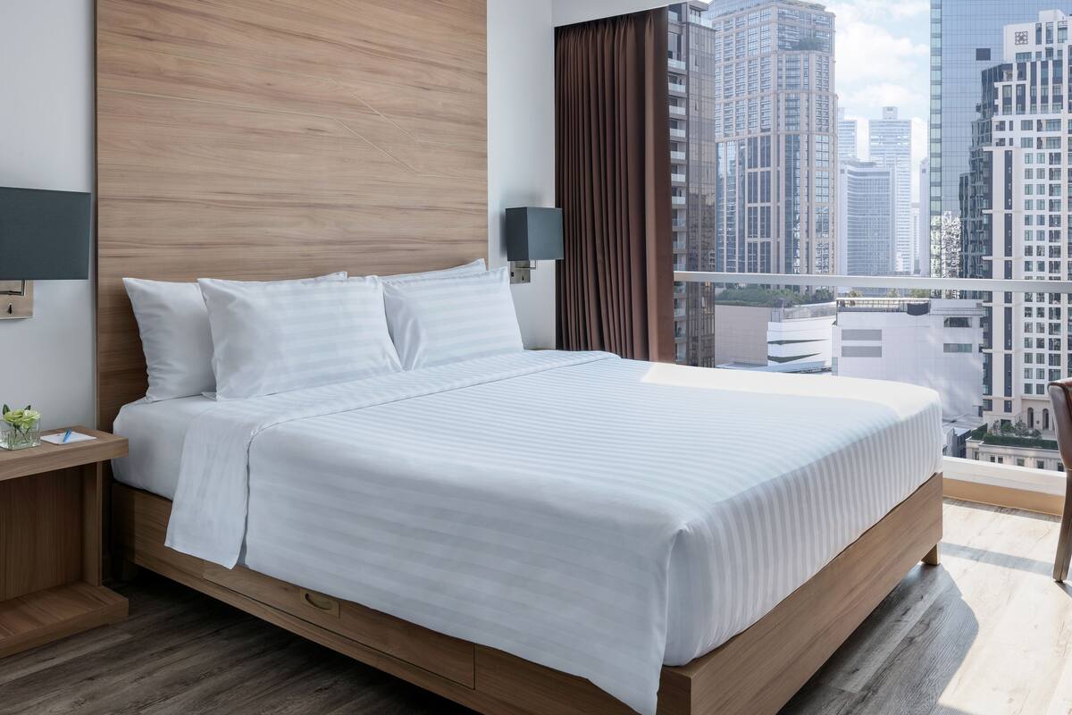 A modern hotel room with a large bed, wooden headboard, two bedside tables with lamps, a small table with a vase, and a view of tall buildings.