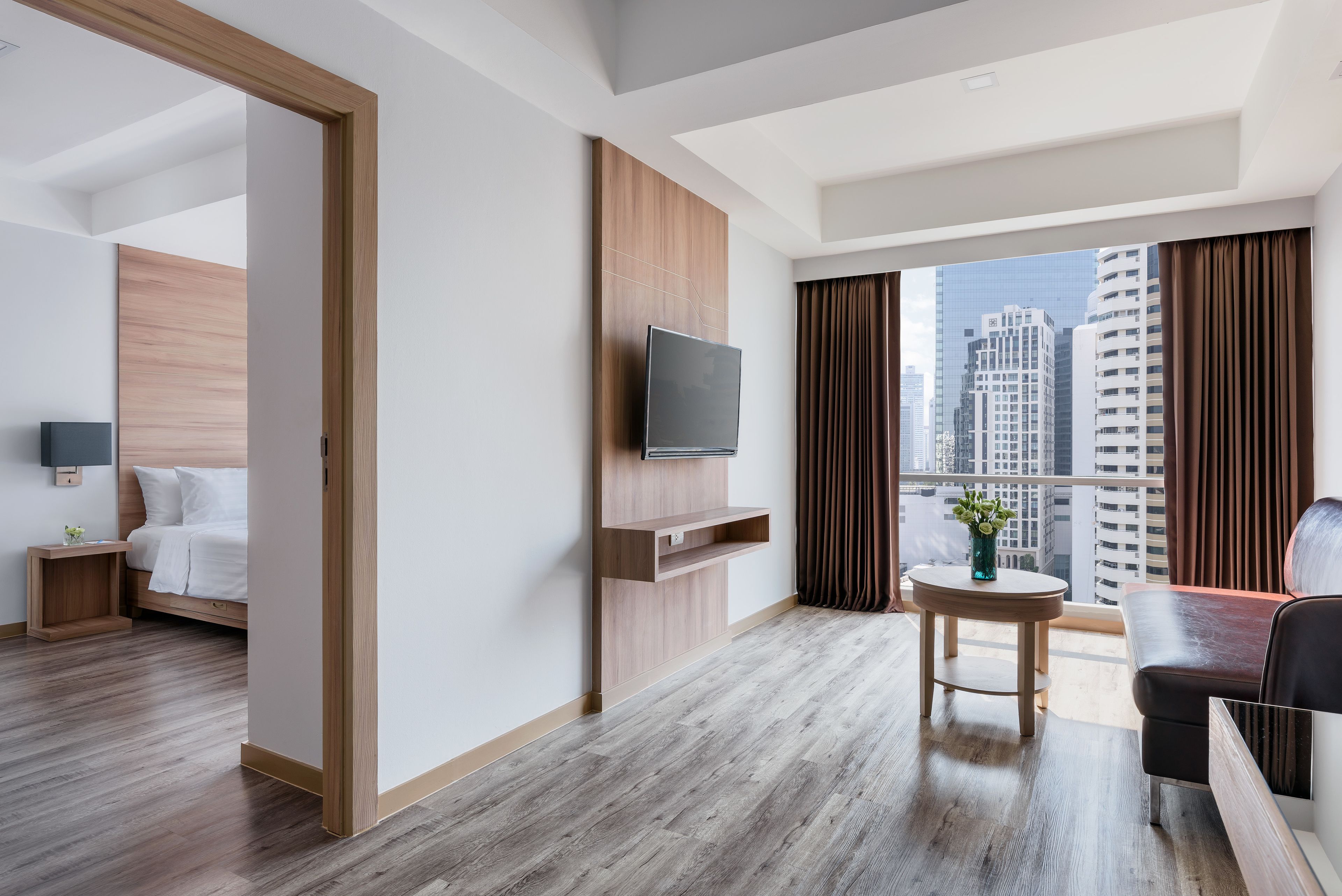 A modern hotel room with a wooden floor, a bed, a TV mounted on the wall, a small table with a plant, and a view of tall buildings through large windows.