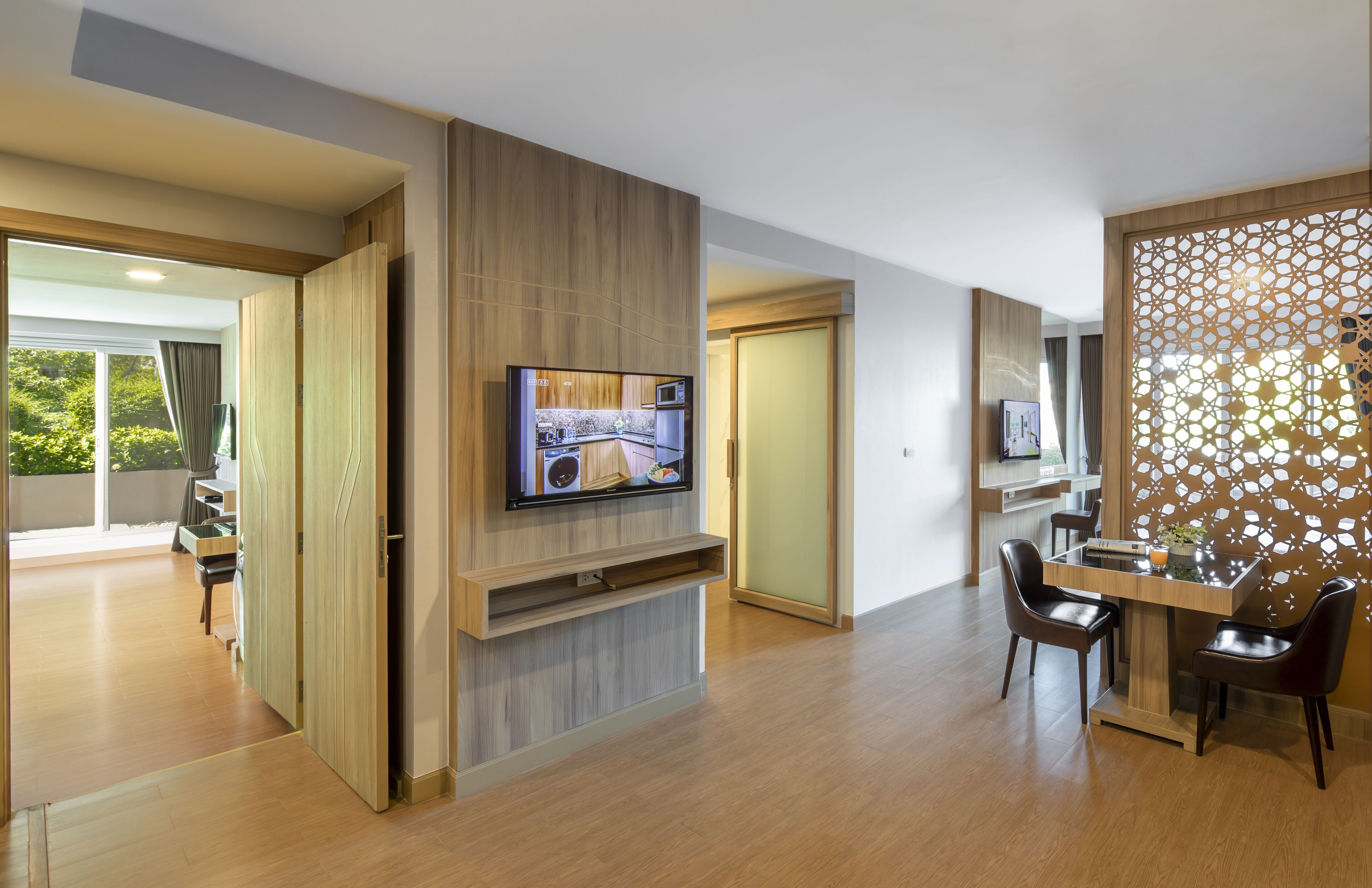 Een moderne woonkamer met een flatscreen televisie aan een houten wand, een eethoek met tafel en stoelen, en uitzicht op de keuken door een open deur.