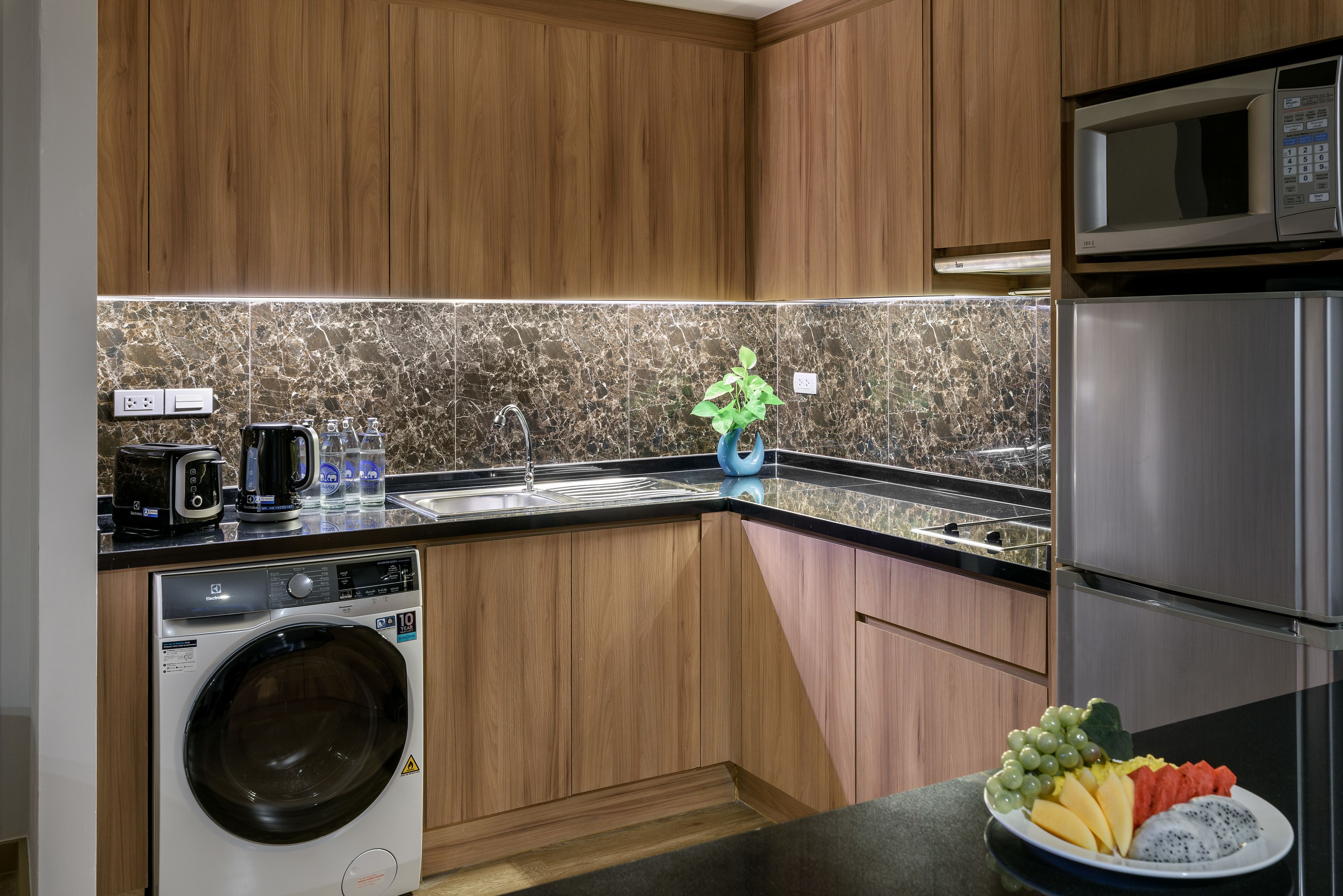 A modern kitchen with wooden cabinets, a built-in washing machine, a stainless steel refrigerator, and a countertop with a fruit bowl.