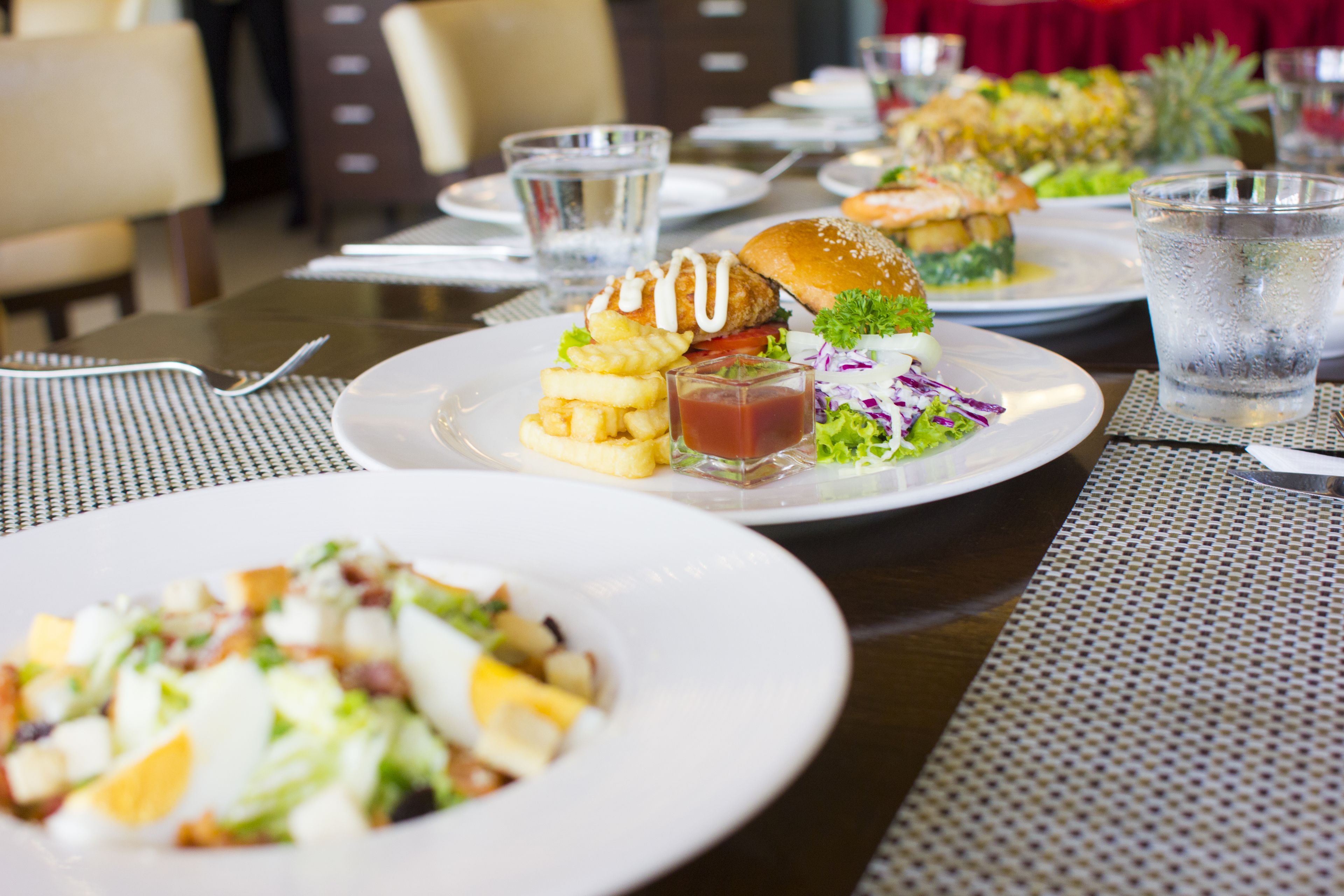 Un restaurante con varios platos de comida, incluyendo hamburguesas, ensaladas y acompañamientos, dispuestos sobre una mesa con mantel a cuadros.