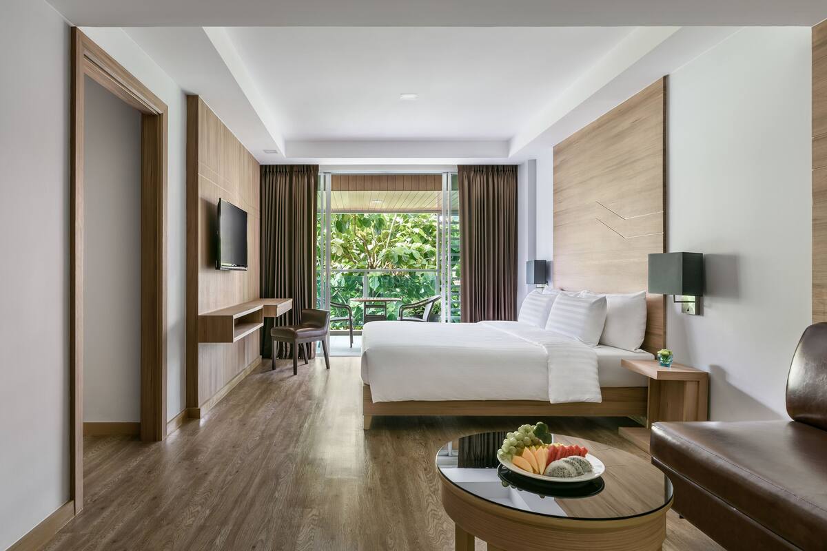 A modern hotel room with a large bed, a desk, a TV, and a view of greenery.